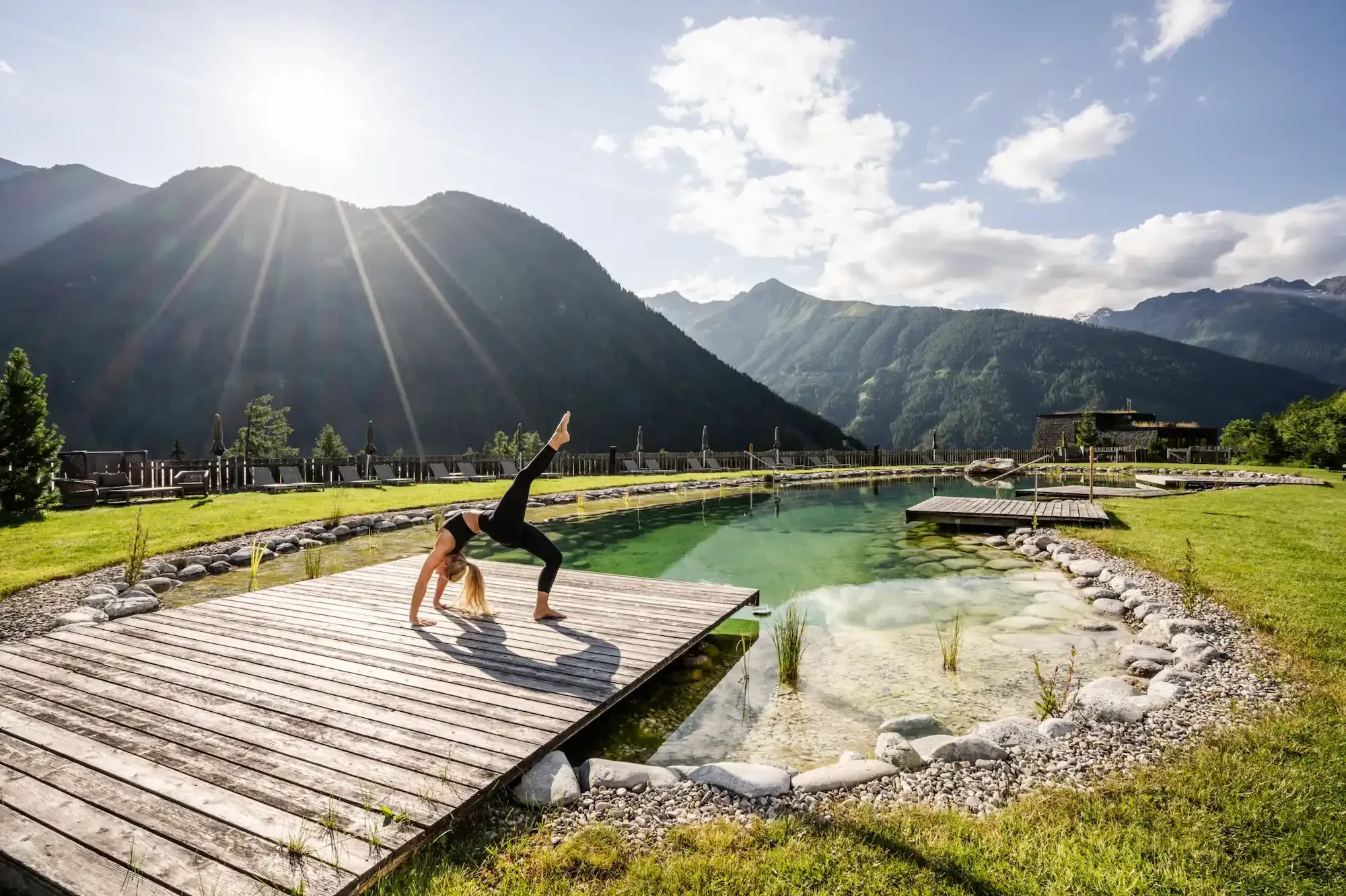 Frau in der Rad-Haltung auf einem Holzsteg am Naturbadeteich vor sonnigem Bergpanorama – Yoga im Gradonna Mountain Resort in Osttirol